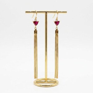 May include: A pair of gold-colored dangle earrings with a triangular, ruby-colored gemstone and long, gold-colored fringe. The earrings are displayed on a gold-colored jewelry stand against a white background.