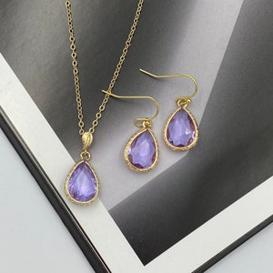 Può includere: Un set di collana e orecchini in oro con cristalli viola a forma di goccia. La collana ha una catena delicata e un ciondolo con un design dettagliato. Gli orecchini sono di stile pendente con un design simile.
