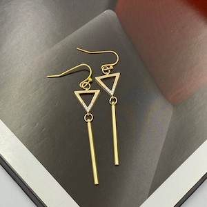 May include: Gold-colored dangle earrings. Each earring features a triangle with a line of small, clear stones along one side, and a long, straight bar hanging below. The earrings have a hook closure.