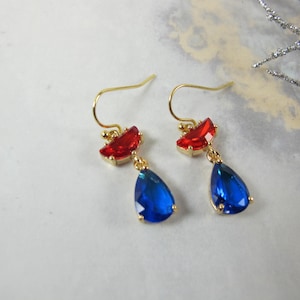 May include: A pair of gold-coloured earrings with red and blue gemstones. Each earring has a red, fan-shaped stone at the top and a teardrop-shaped blue stone hanging below. The earrings are designed with a hook style.