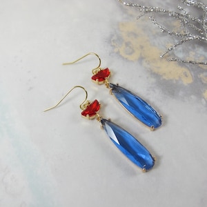 May include: A pair of gold-coloured drop earrings. Each earring has a small red accent and a large, faceted, teardrop-shaped blue crystal pendant. The earrings are presented on a light-coloured surface.