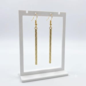 May include: A pair of gold-toned drop earrings featuring long, slender rectangular bars with a textured, hammered surface. The earrings are suspended from gold-toned fishhook ear wires, creating a simple, elongated vertical design.