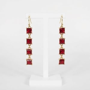 May include: A pair of gold-tone drop earrings, each featuring four square-cut, deep red faceted stones arranged in a vertical line. The earrings are suspended from gold-tone hooks and showcase a polished metal finish with vibrant red gems.