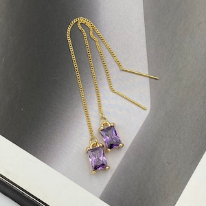May include: A pair of gold-tone threader earrings featuring rectangular purple gemstones. Each earring consists of a delicate gold chain that connects to a prong-set, faceted purple stone at one end and a straight gold post at the other end.