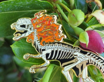 May include: Gold-tone enamel pin of a skeletal dinosaur with a fiery mane. The dinosaur is depicted in a running pose with its tail curled. The pin is set against a background of green leaves and white flowers.
