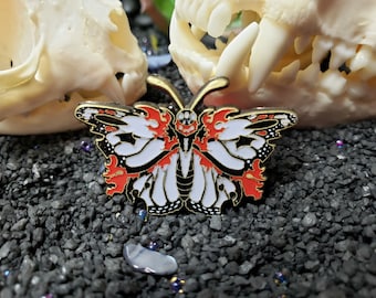 以下が含まれることがあります： 赤色のアクセントが付いた黒と白の蝶のピン。蝶は羽に頭蓋骨のようなデザインが施されています。ピンは黒い背景の上にあります。
