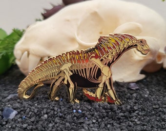Puede incluir: Pin de esmalte dorado de un esqueleto de dinosaurio con acentos rojos y amarillos. El dinosaurio está de pie sobre sus patas traseras con la cola enroscada alrededor de su cuerpo.