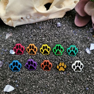 May include: A set of ten colorful paw print charms with black paw prints inside. The charms are arranged in a rainbow pattern on a dark gray background.