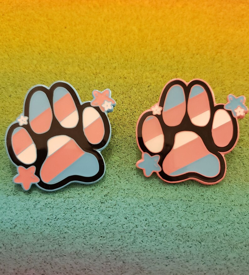 May include: Two paw print pins featuring a transgender pride flag design. The paw prints are black with blue, pink and white stripes. Each paw print has five white stars around it.