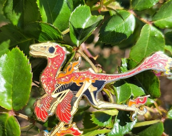 May include: A red and gold enamel pin of a skeletal dinosaur with flames coming from its feet and wings. The dinosaur is in a running pose with its mouth open.