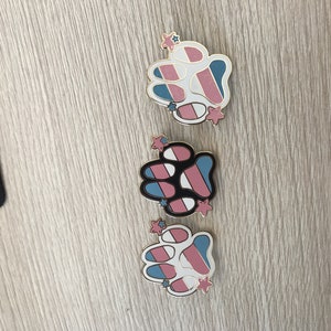 May include: Three paw print pins featuring a transgender pride flag design. Each pin has a white paw print with blue, pink and white stripes. The pins have gold accents and small stars.