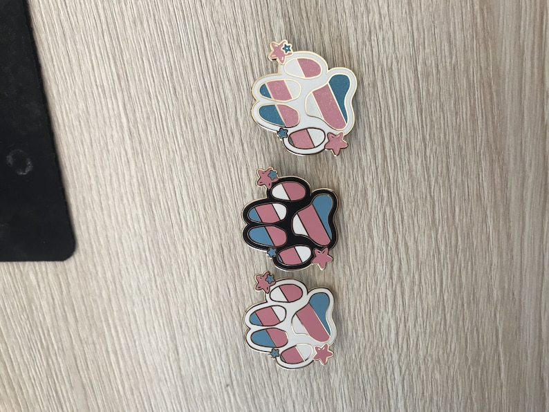 May include: Three paw print pins featuring a transgender pride flag design. Each pin has a white paw print with blue, pink and white stripes. The pins have gold accents and small stars.