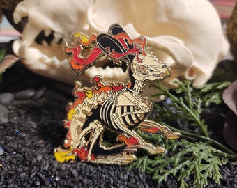 Puede incluir: Un pin de esmalte dorado que representa una criatura esquelética con llamas. El pin presenta un diseño detallado del esqueleto de la criatura, con detalles en negro y crema. Las llamas en tonos rojos, naranjas y amarillos emanan del cuerpo de la criatura.