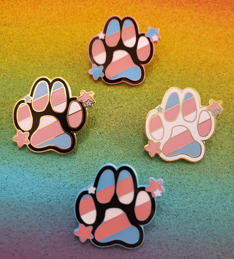 May include: Four enamel pins featuring a paw print design with the transgender pride flag colours. Each pin has a black outline and is decorated with small white stars.