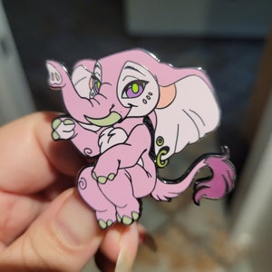 May include: A pink elephant enamel pin with green eyes and a light pink tail. The elephant is wearing a gold charm with a green gem.