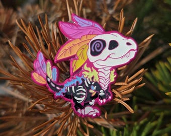 May include: A colorful enamel pin featuring a stylized dragon with a skull-like head and skeleton details. The dragon has pink, yellow, and blue accents, with a vibrant, multi-colored tail and feather details. The pin is set against a blurred background of pine needles.