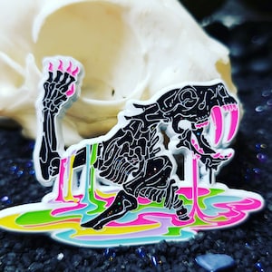 Puede incluir: Un pin de esmalte negro de una criatura esquelética con dientes rosados que gotea limo de color arcoíris. La criatura está de pie sobre un fondo de color arcoíris.