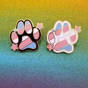 May include: Two enamel pins shaped like a paw print with a transgender pride flag design. The pins are gold-toned and have a white background with pink, blue, and light blue stripes. One pin has a black outline and the other has a white outline. Both pins have small gold stars on the side.