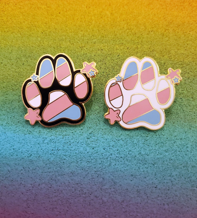 May include: Two enamel pins shaped like a paw print with a transgender pride flag design. The pins are gold-toned and have a white background with pink, blue, and light blue stripes. One pin has a black outline and the other has a white outline. Both pins have small gold stars on the side.