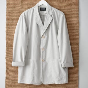 May include: A light gray DKNY blazer with a notched lapel, three buttons, and two front pockets. The blazer is hanging on a white hanger against a textured brown background.