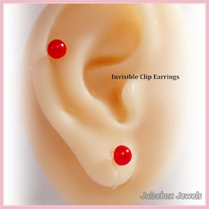 May include: Close-up of a light-toned ear with two small, bright red, round earrings. The text "Invisible Clip Earrings" is visible. The earrings are positioned on the earlobe and upper ear.