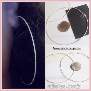 May include: A pair of large, silver hoop earrings with a textured finish. The earrings are shown on a model's ear and also laid flat next to a US quarter for size comparison. The earrings are labeled "Invisible clip on" and "Juicebox Jewels".