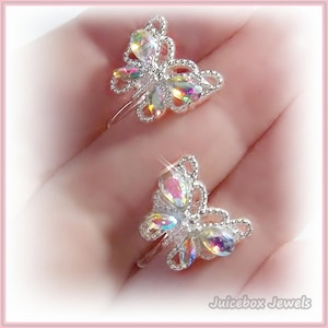 May include: Two silver butterfly rings with iridescent crystal accents. The rings are on a finger and the background is a light pink.