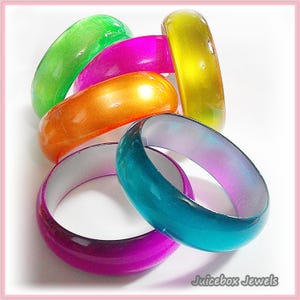 May include: A collection of colorful bangles in shades of green, pink, orange, yellow, teal, and purple. The glossy, rounded bracelets are stacked together, showcasing a variety of vibrant hues. The image is set against a white background.