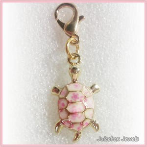 May include: A gold-colored charm featuring a turtle design. The turtle's shell is white with pink floral accents. The charm has a lobster clasp for easy attachment to a bracelet or necklace. The charm is from Juicebox Jewels.