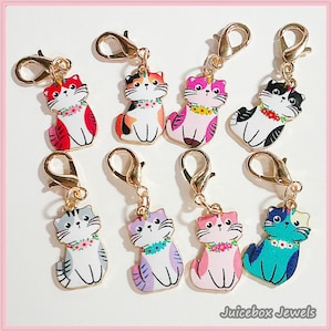 May include: A collection of colorful cat-shaped charms with gold-colored clasps. Each charm features a unique color scheme, including red, orange, pink, black, gray, purple, and teal, with floral collars. The charms are designed with a cartoon-like aesthetic.