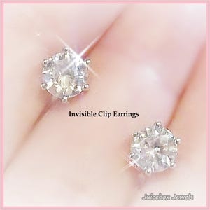 May include: Two clear, round, clip-on earrings with a sparkling, faceted design. The earrings are set against a soft, pink background. Text on the image reads "Invisible Clip Earrings" and "Juicebox Jewels."