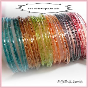 May include: A collection of colorful, translucent bangles in shades of teal, green, orange, and red. The bangles are stacked together, showcasing their varying patterns and textures. The text "Sold in Set of 5 pcs per color" is visible.