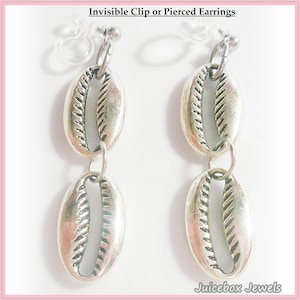 May include: Silver-toned earrings featuring a double cowrie shell design. The earrings are connected by small hoops and have a post for pierced ears or a clip-on option. The text "Invisible Clip or Pierced Earrings" is at the top.