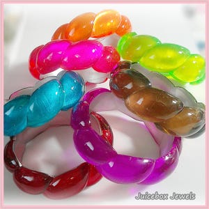 May include: A collection of colorful, translucent bracelets in shades of orange, pink, green, teal, brown, and purple. Each bracelet features a unique, textured design. The bracelets are displayed on a white surface, with the text "Juicebox Jewels" in the bottom right corner.