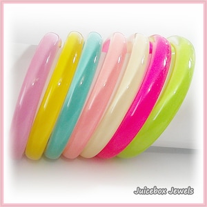 May include: A collection of colorful bangles in shades of pink, yellow, teal, peach, cream, hot pink, and lime green. The bangles are arranged in a diagonal row, showcasing their smooth, glossy surfaces. The image is framed with a pink border.