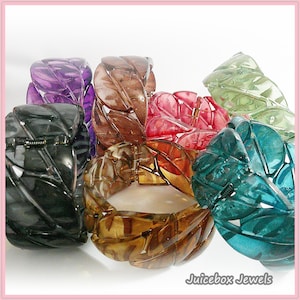 May include: A collection of colorful leaf-shaped bracelets in various shades, including purple, brown, red, green, and teal. Each bracelet features a hinged clasp and is made of a translucent material. The bracelets are arranged in a group, showcasing their diverse colors and designs.