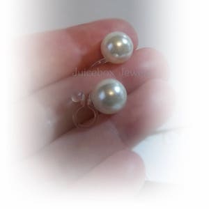 May include: A pair of white pearl clip-on earrings. The earrings feature a simple design with a single pearl on each earring. The pearls are a creamy white colour and have a smooth, glossy finish. The earrings are held in place by a clear plastic clip.