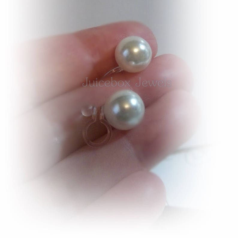 May include: A pair of white pearl clip-on earrings. The earrings feature a simple design with a single pearl on each earring. The pearls are a creamy white colour and have a smooth, glossy finish. The earrings are held in place by a clear plastic clip.