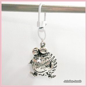 May include: A silver-colored frog charm with large eyes and a textured body, attached to a white clip. The charm is suspended from a metal bar. The text "Juicebox Jewels" is in the bottom right corner.