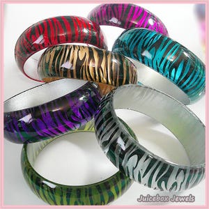 May include: A collection of colorful bangles with a zebra stripe pattern. The bracelets are in various colors, including red, gold, purple, green, blue, and silver. The inside of the bangles is white or silver, and the text "Juicebox Jewels" is visible.