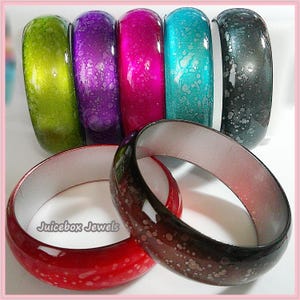 May include: A collection of seven thick bangle bracelets featuring a glossy finish with speckled patterns in lime green, purple, magenta, turquoise, dark gray, red, and a dark brown-to-red gradient. The text Juicebox Jewels is visible.