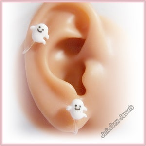 May include: A pair of white ghost-shaped earrings with a smiling face. The earrings are made of plastic and are attached to a clear plastic post.