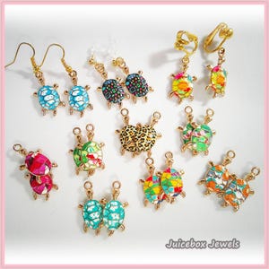 May include: A collection of gold-toned turtle-shaped jewelry pieces, including earrings with hooks, clip-on earrings, and charms. Each turtle features a colorful, patterned shell design in various hues like blue, pink, green, orange, and leopard print.