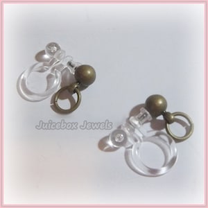 May include: Two pairs of clear plastic earring backs with a small gold-toned ball and a small gold-toned loop.  Juicebox Jewels.