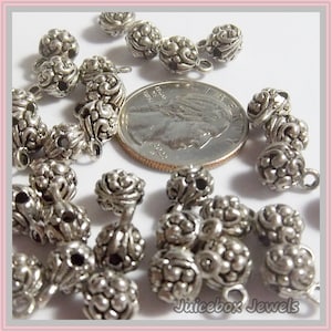 May include: A close-up of silver metal beads with intricate designs. The beads have a loop on one side and a small hole on the other side. The beads are arranged in a random pattern on a white surface. The beads are approximately 1/2 inch in diameter.
