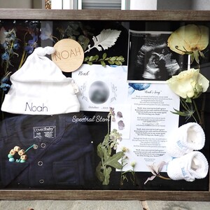 May include: A shadow box with a white baby hat, a blue onesie, a wooden circle with the name "Noah" on it, a small wooden bead necklace, a white crocheted baby bootie, a dried flower arrangement, a poem titled "Noah's Song", and a black and white ultrasound image.