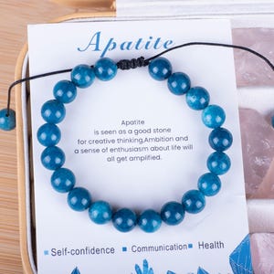 Puede incluir: Un brazalete de piedra de apatita azul con un cordón negro. El brazalete está sobre una tarjeta blanca con el texto "Apatite is seen as a good stone for creative thinking Ambition and a sense of enthusiasm about life will all get amplified." y las palabras "Self-confidence Communication Health".
