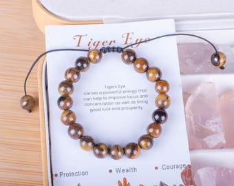 May include: A brown tiger's eye beaded bracelet with a black cord closure. The bracelet is displayed on a white card with the text "Tiger Eye" and a description of the stone's properties. The card also lists the benefits of the stone, including protection, wealth, and courage.