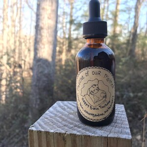 K&ouml;nnte beinhalten: Eine dunkelbraune Glasflasche mit Tropfer und einem Etikett mit der Aufschrift "Medicine of Our Mother Happy Brain Tincture Balm Ashwagandha, Motherwort". Die Flasche steht auf einer Holzoberfl&auml;che.