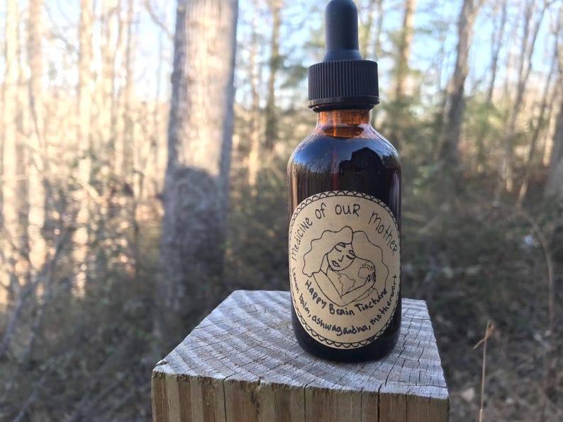 K&ouml;nnte beinhalten: Eine dunkelbraune Glasflasche mit Tropfer und einem Etikett mit der Aufschrift "Medicine of Our Mother Happy Brain Tincture Balm Ashwagandha, Motherwort". Die Flasche steht auf einer Holzoberfl&auml;che.